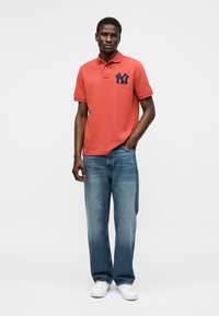 Homme portant un polo corail avec un logo marine, un jean bleu et des baskets blanches, debout avec une main dans la poche sur un fond uni.