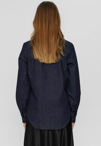 Mörk denimskjorta med långa ärmar, rundad nederkant och en enkel krage. Har sömnadsdetaljer på ärmsluten och axlarna, slät textur.