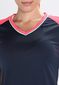 Námořnické sportovní tričko s výstřihem do V, které má růžové a tyrkysové akcenty na límci a ramenou. Hladká textura, krátké rukávy.