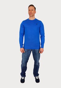 Modra pletena jopica z dolgimi rokavi, okroglo izrezom in rebrastim robom. Nošena z modrimi kavbojkami in črnimi supergami. Ima majhen logo na prsih.