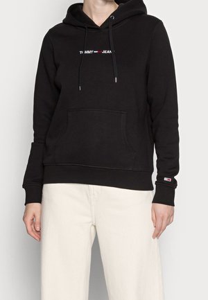 Sudadera negra con bolsillo frontal y logo de "Tommy Jeans" en el pecho y la manga, combinada con pantalones color crema en una persona de pie.