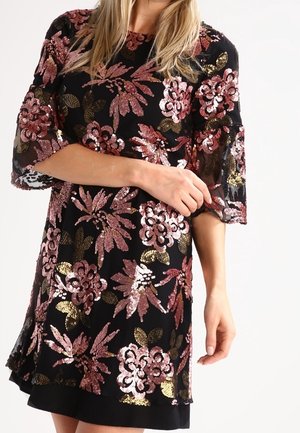 Zwarte jurk met roze en gouden pailletten bloemenpatronen, driekwart doorschijnende mouwen, gedragen door een persoon die de mouw aanpast.