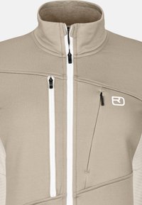 Beige Fleecejacke mit hohem Kragen, ausgestattet mit zwei vorderen Reißverschlusstaschen und einer Brusttasche. Weiße Reißverschlüsse und Logo betonen das Design.