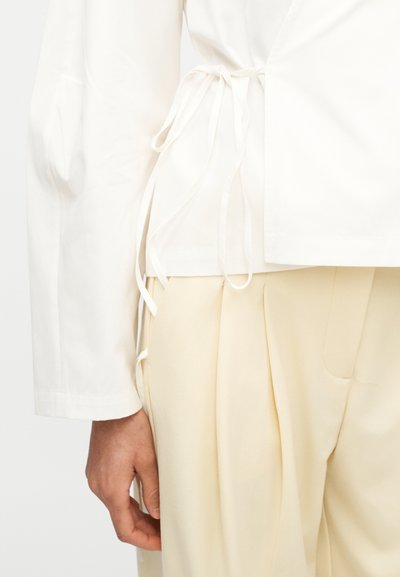 Κοντινό πλάνο ενός ατόμου που φοράει κρεμ πλισέ παντελόνια και λευκό πουκάμισο τύπου wrap δεμένο στη μέση με κρεμασμένες υφαντικές κορδέλες.