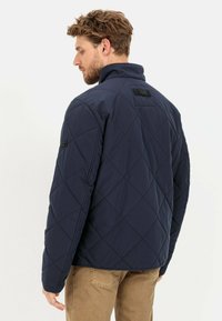 Navy gesteppte Jacke mit hohem Kragen, diagonalen Steppnähten und schwarzen Hardware-Akzenten an den Schultern und im Rücken.