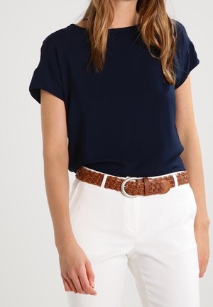 Femme portant un haut bleu marine à manches courtes rentré dans un pantalon blanc avec une ceinture en cuir tressé marron avec une boucle en argent.