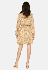 Eine Frau steht mit dem Rücken zur Kamera, trägt ein langärmeliges Kleid mit beige-weißer Zebramusterung, das an der Taille tailliert ist, und nacktfarbene Blockabsatzschuhe.