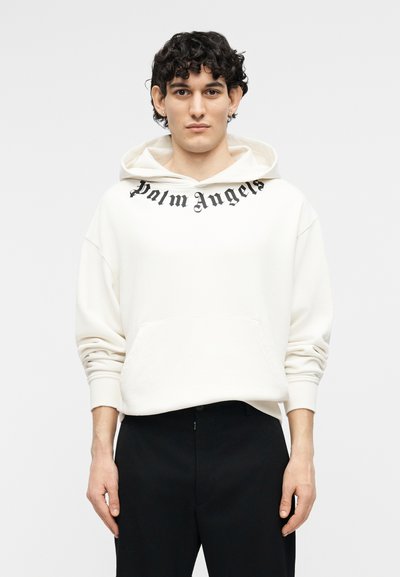 Jeune homme aux cheveux bouclés portant un sweat à capuche blanc Palm Angels et un pantalon noir, debout devant un fond clair uni.