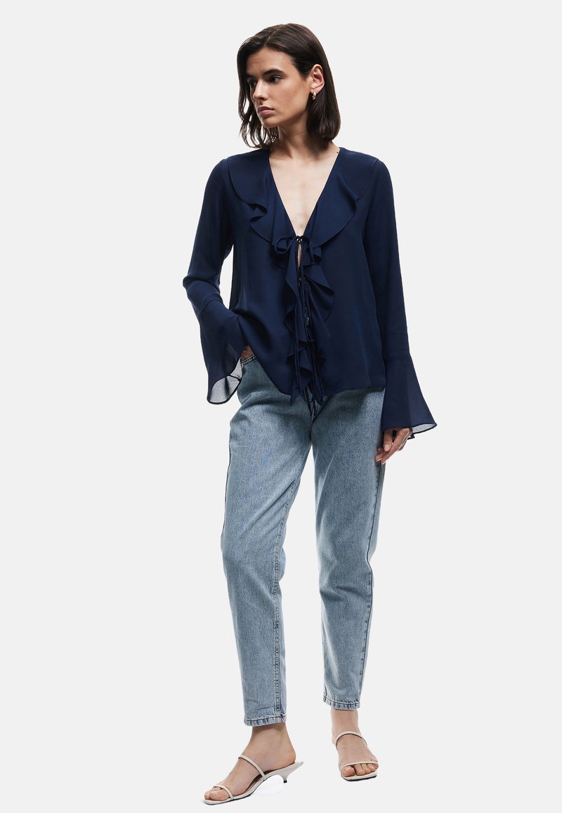 Blusa navy con frente con volantes, escote en V profundo y puños acampanados, combinada con jeans de mezclilla azul claro y sandalias minimalistas.