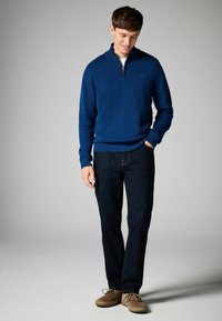Marineblauer Pullover mit Viertelreißverschluss aus strukturierter Stoff, kombiniert mit dunklen Jeans und hellbraunen Schuhen, präsentiert ein lässiges Outfit.