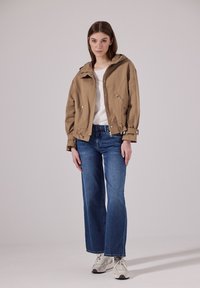 Vaaleanruskea, hupullinen, lyhyt takki, jossa on nepparit ja laatikkomainen istuvuus, paritettu sinisten leveiden farkkujen ja valkoisten tennareiden kanssa.