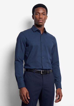 Mann trägt ein eng anliegendes dunkelblaues Hemd mit Knöpfen, das in marineblaue Hosen mit schwarzem Gürtel gesteckt ist, und steht vor einem schlichten weißen Hintergrund.