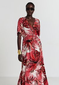 Femme portant une robe d'été à motifs tropicaux rouge et blanc, lunettes de soleil sombres et large bracelet en or, se tenant devant un fond clair.