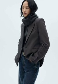 Grå rutmönstrad blazer med spetsiga slag, bärs över en mörk polotröja. Kombinerat med blå högmidjade jeans, har outfiten en texturerad scarf.