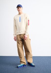 Μπεζ φούτερ με μπλε ετικέτα "ALPHA" και κόκκινες λεπτομέρειες, συνδυασμένο με χαλαρές καφέ cargo �παντελόνες και μαύρα με μπλε μοτίβο sneakers.