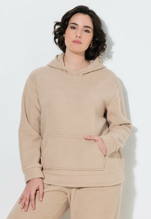 Ung person med kort krøllet mørkt hår, iført en beige hættetrøje og matchende bukser, stående mod en hvid baggrund.