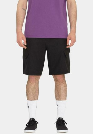 Pantalones cortos de carga negros con bolsillos laterales, combinados con una camiseta morada y calcetines blancos con un logo. El conjunto se completa con zapatillas negras.