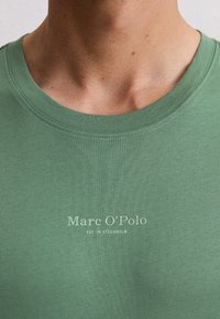 Zbliżenie na osobę w zielonym T-shircie z okrągłym dekoltem, na którym widnieje napis "Marc O’Polo EST. W Sztokholmie".