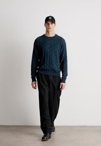 Mørkeblå teksturert genser med diamantmønster, ribbede mansjetter og midjebånd. Kombinert med løse svarte bukser og en svart caps.