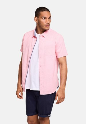 Hombre de pie mirando hacia un lado, vistiendo una camisa de manga corta rosa claro sobre una camiseta blanca y pantalones cortos azul marino, sobre un fondo liso.