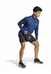 Blauw sportshirt met lange mouwen, zwarte sportshorts, houdt een kettlebell vast, draagt zwarte sportschoenen. Glad materiaal, atletische pasvorm, logo op het shirt.