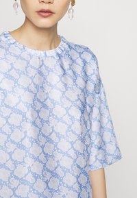 Blusa azzurra chiara con un motivo floreale bianco, maniche corte e scollatura arricciata. Presenta una texture di tessuto liscia e morbida.