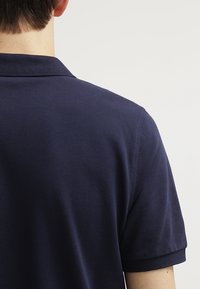 Granatowa koszulka polo widoczna z tyłu, z klasycznym kołnierzykiem i krótkimi rękawami. Materiał jest teksturowany, z delikatnym, tkanym wzorem.