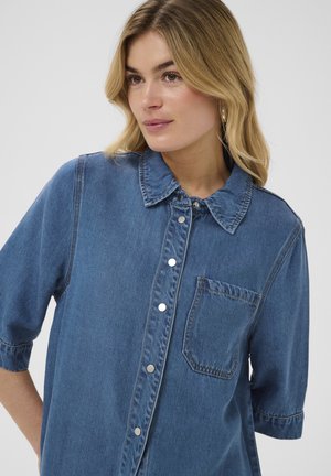 Femme blonde portant une chemise en jean bleu à boutons avec une poche poitrine, regardant légèrement sur le côté sur un fond uni.