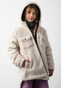 Kremowa polarowa kurtka z kapturem, z przednimi kieszeniami i zapięciem na zamek, noszona na ciemnym swetrze. Faktura jest miękka i puszysta.