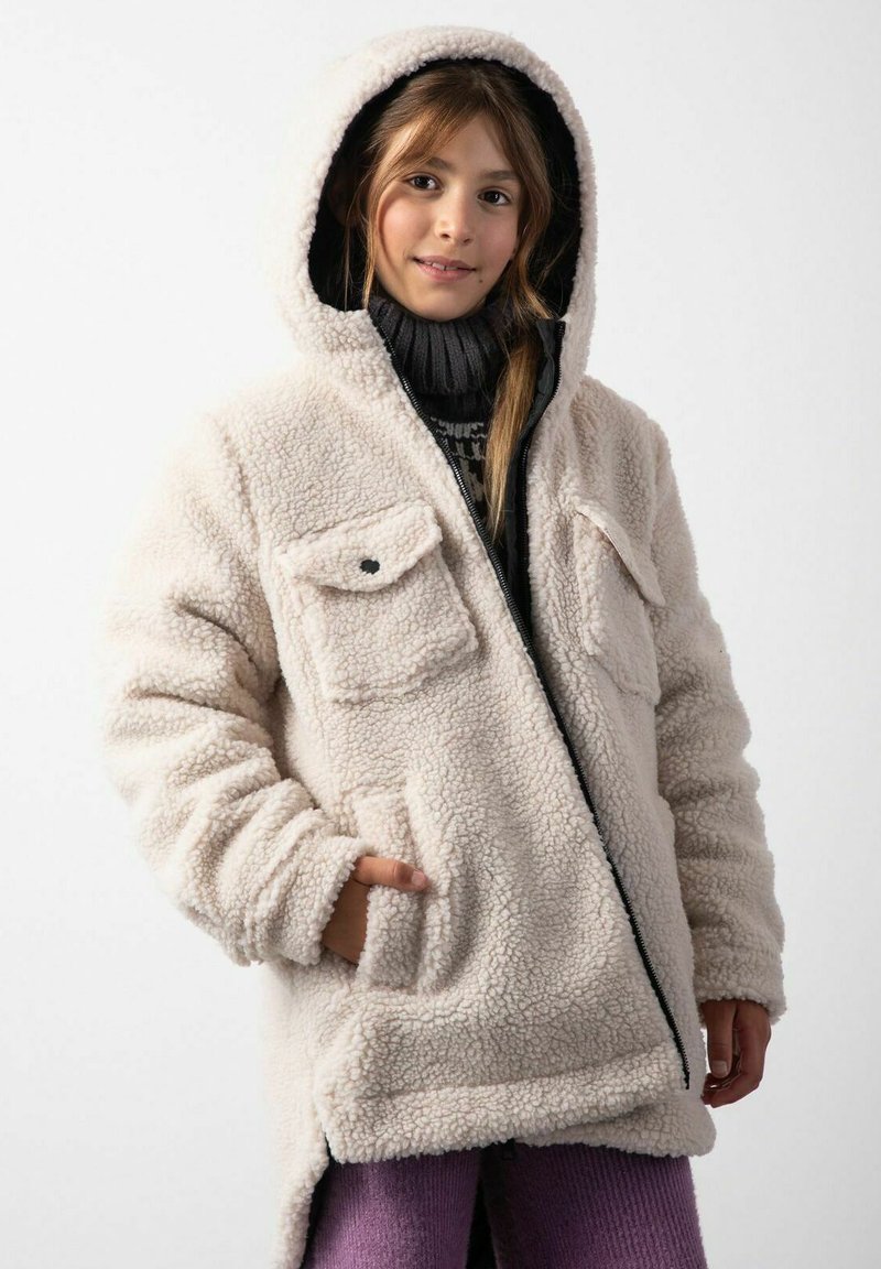 Kremowa polarowa kurtka z kapturem, z przednimi kieszeniami i zapięciem na zamek, noszona na ciemnym swetrze. Faktura jest miękka i puszysta.