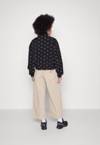 Jachetă neagră cu modele albe, pantaloni cargo bej largi, încălțăminte neagră și șosete albe; ținută casual cu o siluetă relaxată.