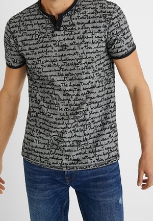 Homme portant un t-shirt henley gris à manches courtes avec un motif de texte en script noir et un bord noir, associé à un jean bleu.