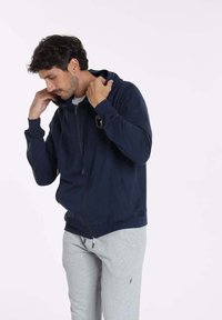 Hoodie zippé bleu marine avec une poche avant, capuche réglable par cordon de serrage et un emblème brodé sur la poitrine. Fabriqué en tissu doux, il possède des poignets côtelés.