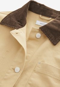 Beige samettijakki, jossa on ruskea samettiolka, hopeiset nepparit ja rintatasku, jossa näkyviä ompelutaitoksia.