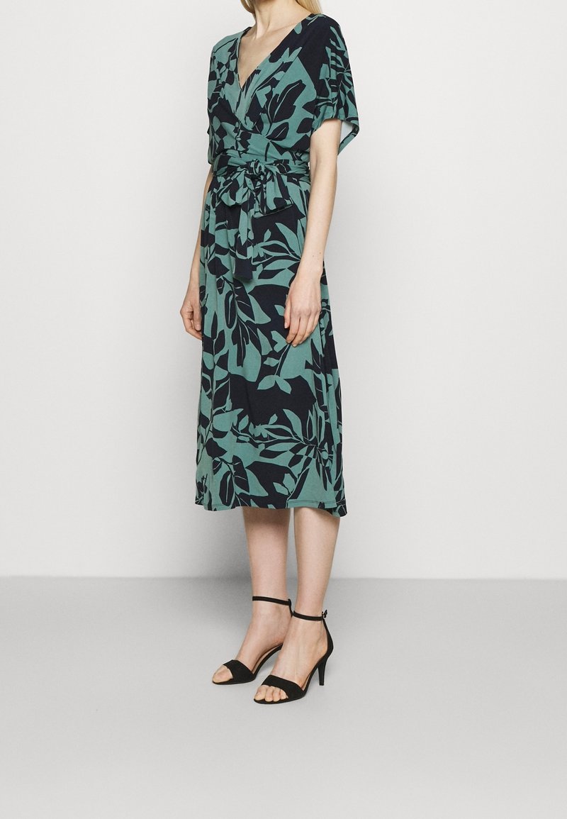 Femme portant une robe verte longueur genou avec un motif de feuilles noires et des manches courtes, associée à des sandales à talons hauts noires avec bride à la cheville.