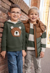 Grön hoodie med björndesign, blåa glasögon, ljusa jeans; brun och grön pufferväst över randig tröja, beige cargobyxor.