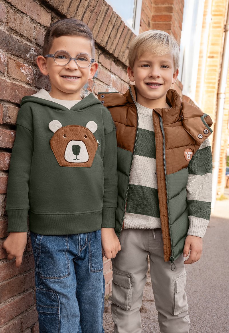 Grön hoodie med björndesign, blåa glasögon, ljusa jeans; brun och grön pufferväst över randig tröja, beige cargobyxor.