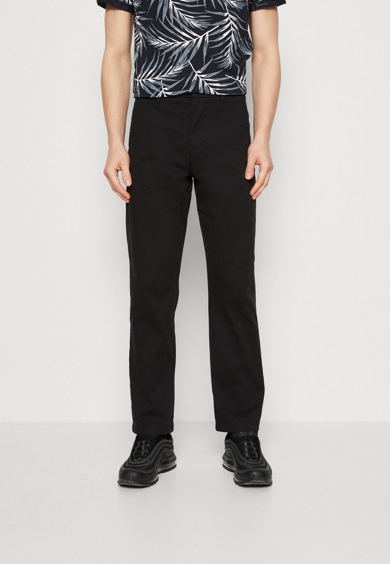 Pantaloni neri con una vestibilità dritta, realizzati in cotone, caratterizzati da una texture liscia e senza motivi visibili. Indossati con una camicia a fantasia e scarpe nere.