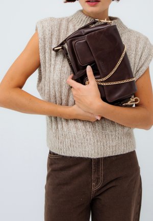 Personne portant un pull sans manches en tricot et un pantalon marron, tenant un sac à main en cuir marron foncé avec une chaîne dorée près de la poitrine.