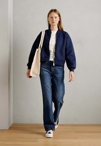 Marine zip-up bomberjakke, hvid skjorte med knapper, løse blå jeans og hvide sneakers. Modellen bærer en beige totebag. Glatte teksturer gennem hele outfittet.
