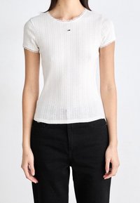 Biała bluzka krótki rękaw o żebrowanej fakturze, z falbaniastym dekoltem i małym logo na piersi. W połączeniu z czarnymi spodniami.