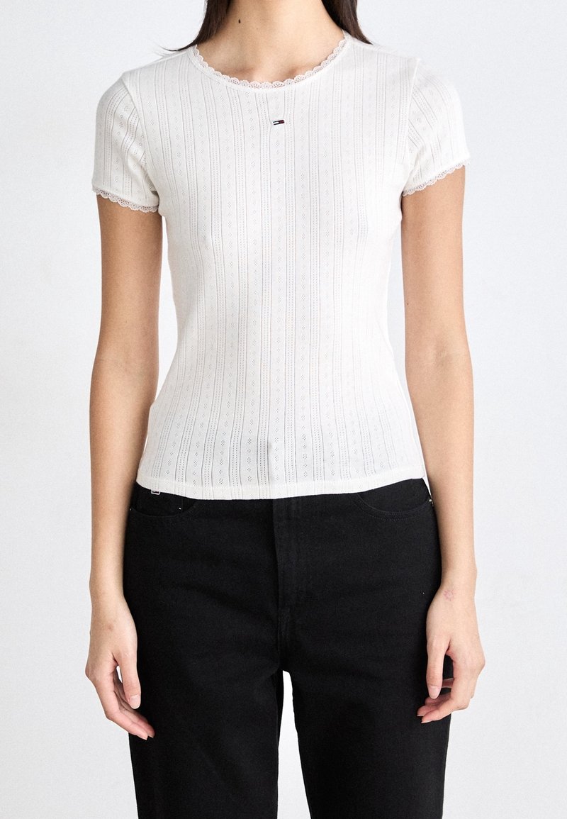Biała bluzka krótki rękaw o żebrowanej fakturze, z falbaniastym dekoltem i małym logo na piersi. W połączeniu z czarnymi spodniami.