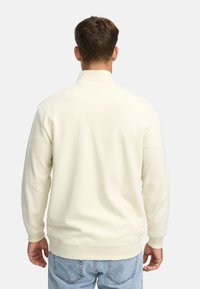 Hombre vestido con un suéter de canalé blanco sucio y jeans azul claro, de pie de espaldas contra un fondo gris claro liso.