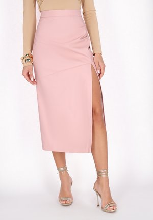 Blush-pink Midi-Rock mit seitlichem Schlitz, glatter Textur, hohem Taillendesign und schrägen Nahtdetails, kombiniert mit goldenen Riemchen-High-Heels.