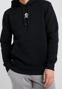 Felpa nera in cotone, con tasca frontale, polsini a coste e logo stampato sul petto. Indossata sopra pantaloni della tuta grigi.