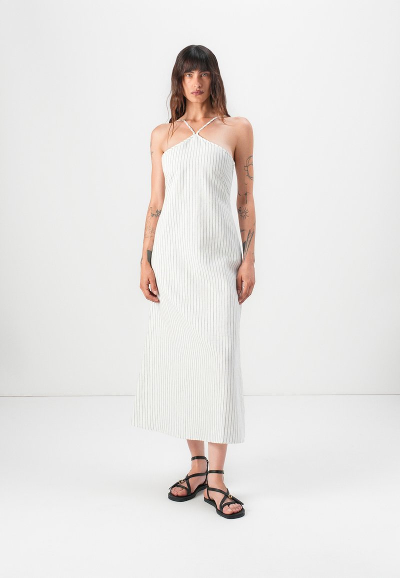 Robe maxi à rayures blanches avec des fines bretelles, un corsage ajusté et une silhouette en A. Le mannequin porte des sandales noires à brides, debout sur un fond blanc.