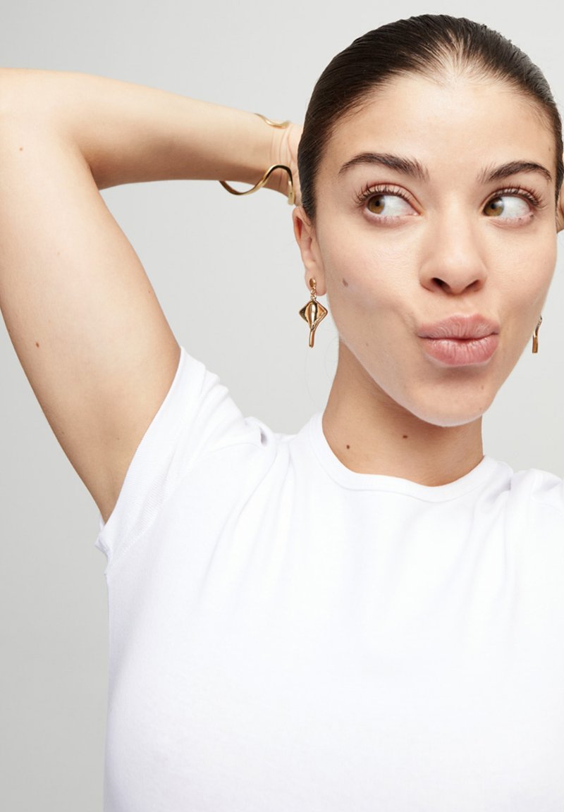 Camiseta blanca de manga corta, tejido liso, con costuras visibles en los hombros. Pendientes dorados en forma de ojos abstractos. Fondo claro.