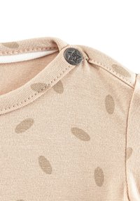 Helles beiges Hemd mit Rundhalsausschnitt, das hellbraune ovale Muster und einen metallischen Sternknopf an der Schulter aufweist. Glatte, weiche Textur.