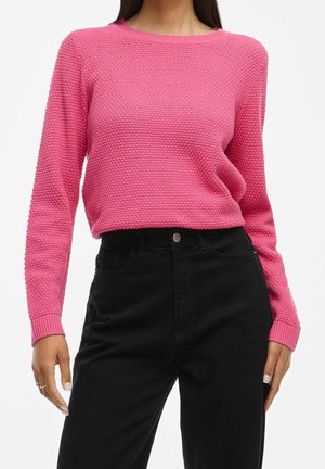 Kvinde iført en struktureret pink langærmet sweater, stoppet ned i sorte bukser med høj talje, stående med hænderne afslappet ved siden.