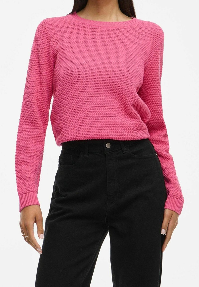Femme portant un pull rose texturé à manches longues, rentré dans un pantalon noir taille haute, debout les mains détendues le long du corps.